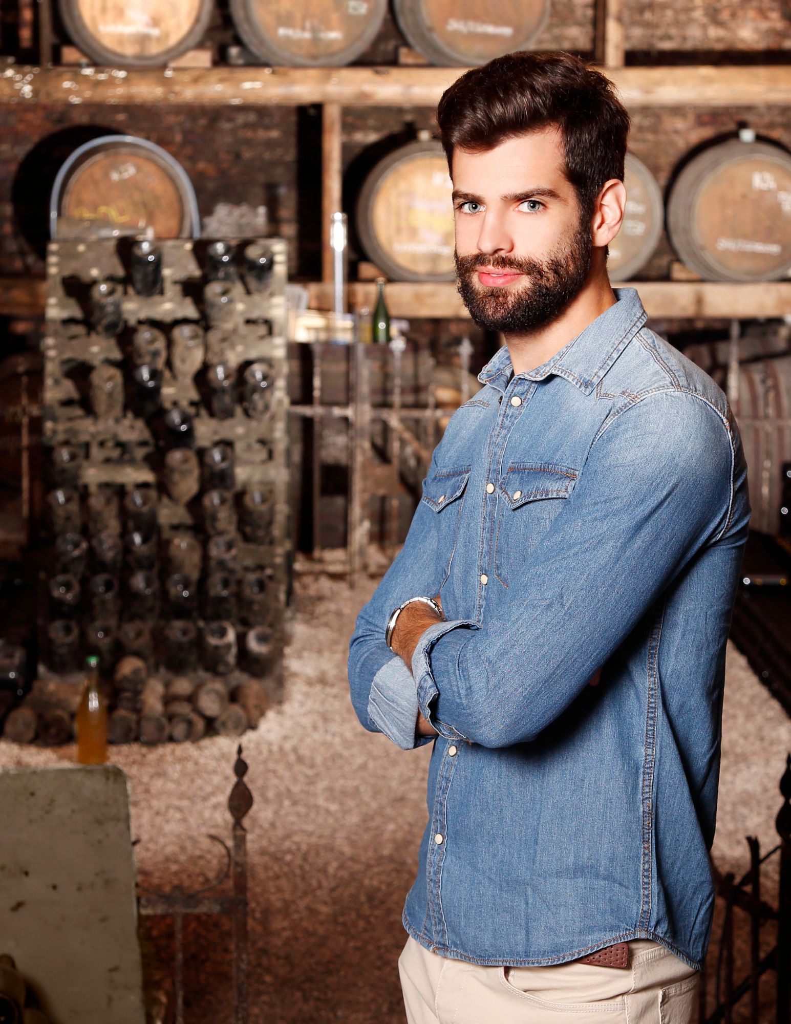 Winery Professional Posing in Barrel Room Confident young man with a beard and denim shirt stands with arms crossed in a wine cellar, surrounded by stacked wine barrels and vintage bottles—symbolizing leadership and expertise in the wine industry.