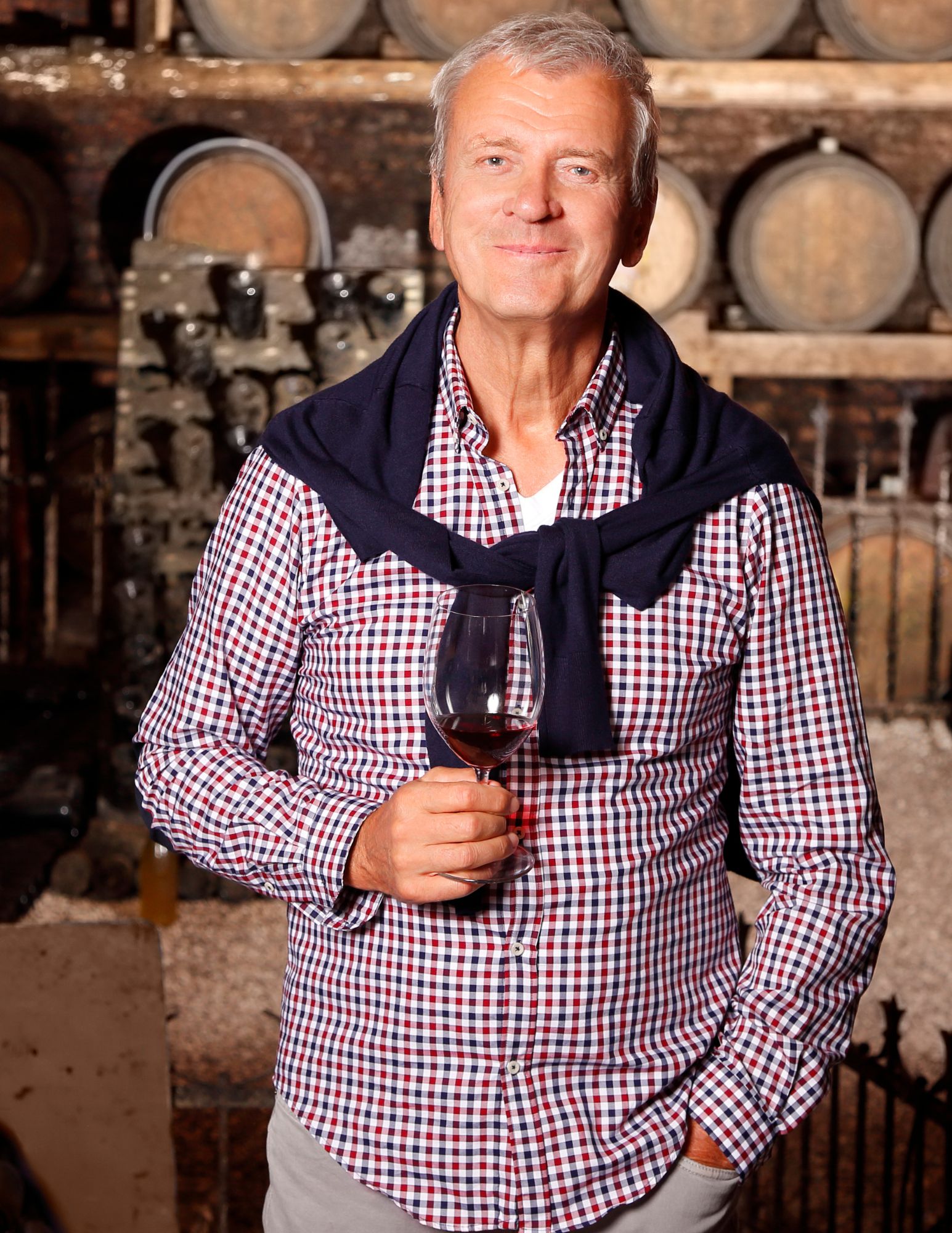 Senior Wine Executive Enjoying a Glass in the Cellar Smiling older man in a checkered shirt and sweater holds a glass of red wine in a winery cellar, representing experience, leadership, and appreciation for craftsmanship in the wine industry.